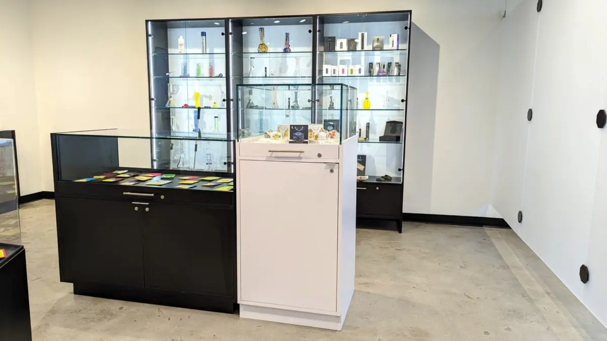 Black floor display case with glass top beside a white pedestal display and tall glass-door cabinets with lit shelves