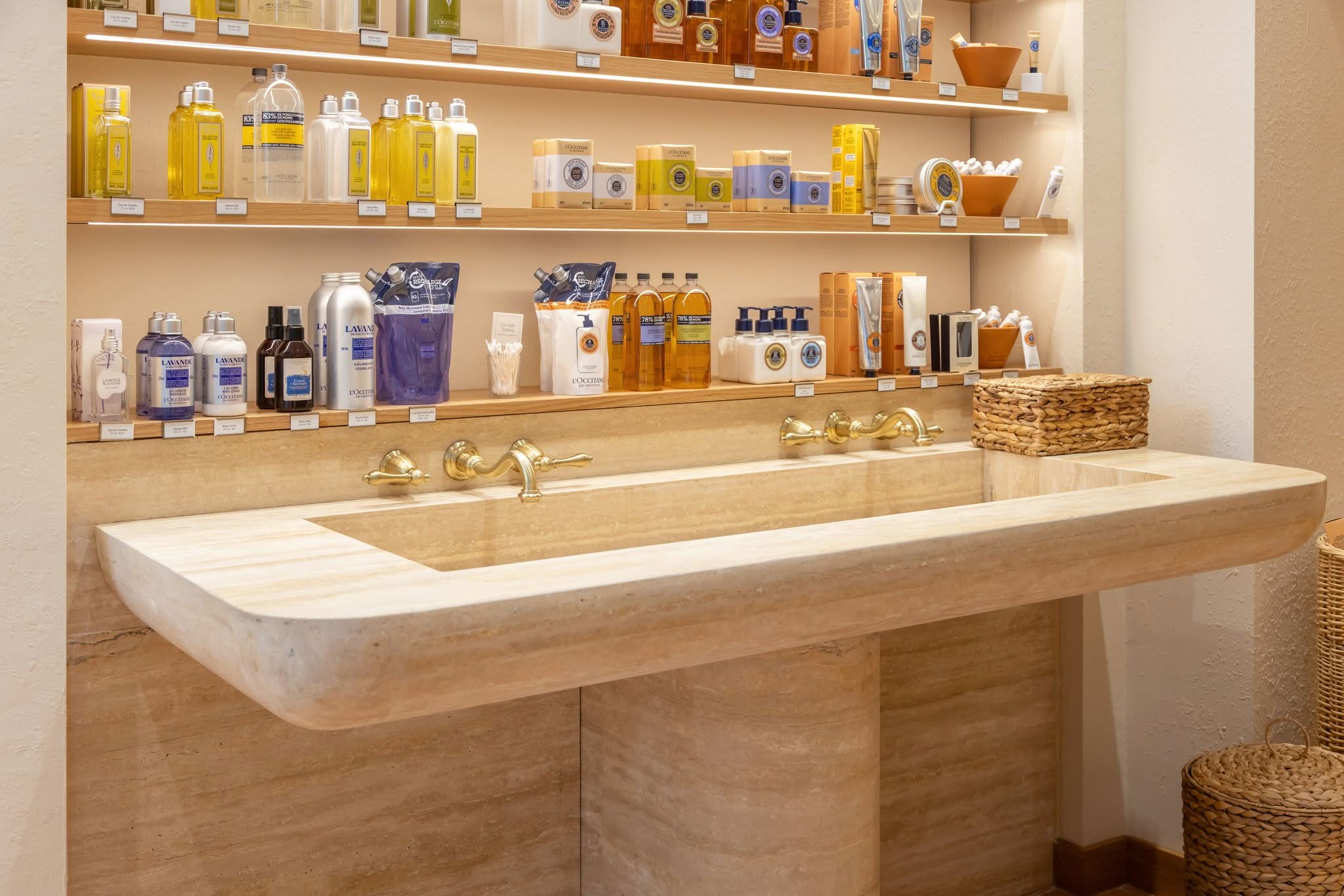 Travertine hand-wash station with carved basin, brass wall-mounted taps and floating oak shelves, fabricated by DNS Industries for L’Occitane Bloor Street Toronto