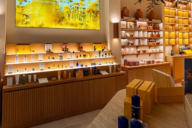 Fluted oak base casework and wall-mounted illuminated shelving running the length of the sales floor, manufactured by DNS Industries for L’Occitane Park Royal, North Vancouver
