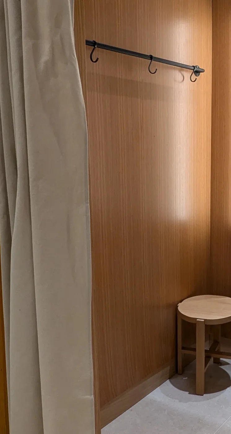 Mango fitting room with wood-veneer partitions, a curtain, a black hook rail and a wooden stool